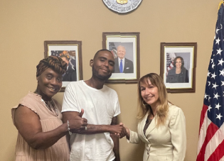 In front of the Department of Labor seal in a federal building, a man is shaking hands with a woman in a suit. Another woman stands beside him, giving a thumbs up to the camera. All are smiling.