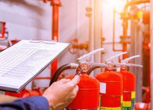 A worker holds a clipboard while inspecting fire extinguishers in an industrial environment