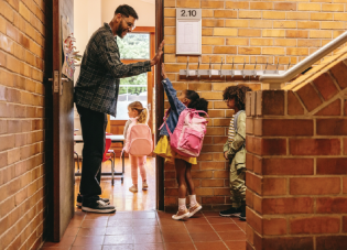 A teacher stands in the doorway of a classroom, giving high fives to young children as they enter.