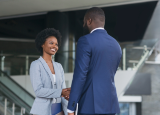 A young woman with a blue blazer smiles while shaking the hand of a man in a blue suit. They are standing in a modern office lobby.
