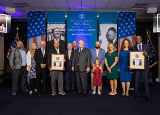 A group of family members and government officials attend a ceremony to induct Sen. Robert Griffin and Howard Jenkins Jr. into the Hall of Honor. Banners behind them have the American flag and black and white photos of both men.
