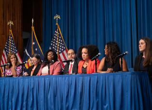 Seven professionally dressed women and men sit on a panel behind a table. American and Labor Department flags are visible on the stage behind them.