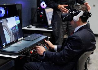 A man in a suit uses a virtual reality headset and aircraft controls as part of a training simulation.