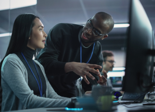 A woman sits at a computer station, engaged in conversation with a man standing next to her.