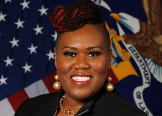 A black woman wearing professional attire smiles for a professional headshot with the American Flag serving as the backdrop. 