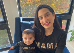 A woman with dark hair sits by a window, holding a baby in her lap.