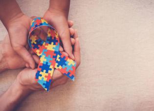 Two sets of hands holding an Autism Awareness Ribbon made of several colorful puzzle pieces