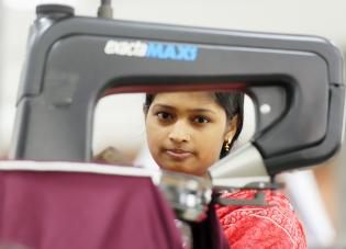 Uma jovem de Bangladesh vestindo roupa vermelha e sentada atrás de uma máquina de costura.