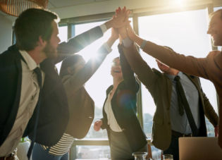 A group of professionals embracing in a high five in an office setting with the sunlight shining in through a large window.