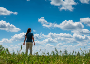 A woman wearing a blue shirt and tan pants stands in an open field and stares up at the sky.