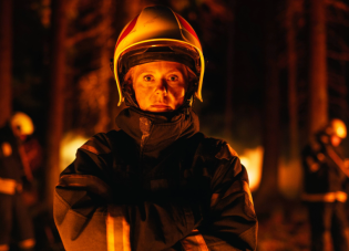 A female firefighter wearing her uniform and protective gear battling a fire in a smoky forest.