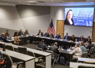 A young woman stands in a large room filled with professionally dressed people seated at tables, and faces a projection screen that reads "Improving UI Customer Experience & Access"