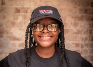 Brianna, a graduate of the STRIVE Future Leaders program, faces the camera smiling and wearing a Cafe Reconcile baseball cap and black t-shirt