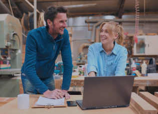 A man and a woman in a workshop are laughing and talking while looking at something on a laptop.