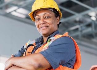 A Black woman wearing protective equipment and a hard hat. She is smiling for a photo with her arms folded.