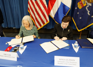 Dalawang babae ang nakaupo sa mesa na lumalagda ng mga dokumento. Nasa likod nila ang mga watawat ng Estados Unidos, Dominican Republic at Departamento ng Paggawa (Department of Labor) ng U.S.