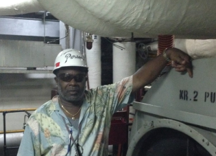 A man stands on a ship wearing a hardhat and protective sunglasses. 