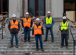 Five men in safety vests and hard hats stand on the crumbling steps of a storm-damaged building