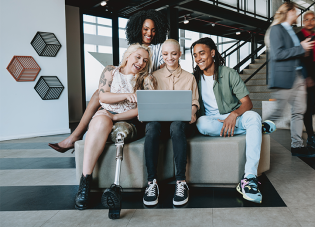 A diverse group of young people – with and without visible disabilities – sit in a building lobby, smiling as they look at a laptop screen.