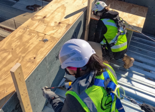 Two female construction employees wearing fall protection gear, including safety helmets, harnesses and other equipment.