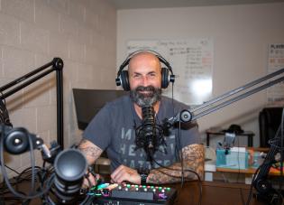 George Nolan sits at a desk wearing headphones with a recording microphone near his mouth. 