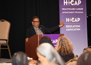 A woman in a dark jacket and glasses speaks at a podium. A banner in the background reads "H-CAP Healthcare Career Advancement Program. H-CAP Education Association."