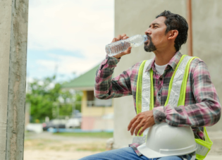 A construction worker sits outside in the shade, holding his hard hat in one hand and drinking water from a clear plastic bottle. 