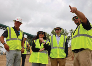 Keith Sonderling and Lori Chavez-DeRemer stand on a construction site with workers, all wearing safety vests and hard hats.