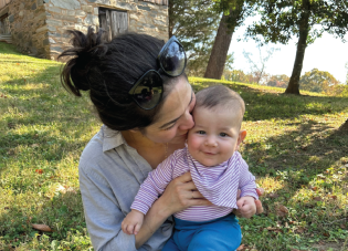 A mother sits on a lawn, holding a smiling baby in her lap and giving it a gentle kiss.