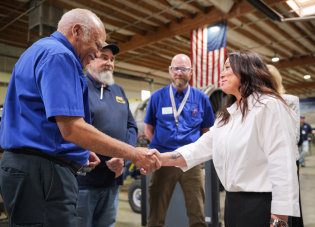 Secretary Lori Chavez-DeRemer shakes the hand of a worker in a blue shirt while several others look on. A large American flag hangs from the ceiling behind them.