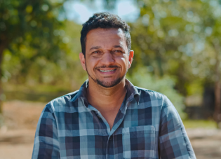 A man wearing a blue checkered button-down collared shirt smiles at the camera while standing in an open field with trees behind him.