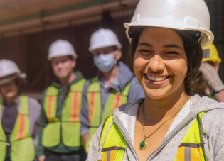 Nakangiti ang isang kabataang babaeng manggagawa na nakasuot ng hard hat, kasama ang ilan pang manggagawang nakatayo sa likuran niya.