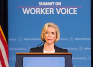 Lilly Ledbetter, an older white woman with short blond hair, is pictured from the shoulders up behind a podium. A blue screen behind her bears the words “Summit on Worker Voice.”