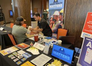 A nurse puts a blood pressure cuff on a miner at a booth with a "Miner Health Matters" banner. Health materials, stickers and other resources are spread out on the table around them.