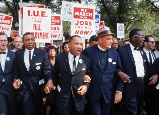 Color image of Martin Luther King, arm in arm with other March leaders, marching in Washington. A crowd behind them holds placards reading "March for jobs for all now" and "IUE AFL-CIO for full employment"