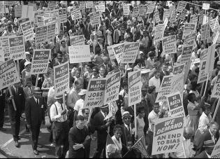 Black and white photo from the Library of Congress shows demonstrators marching in the street holding signs during the March on Washington, 1963.