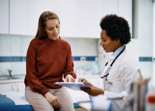 A woman wearing a rust-colored turtleneck and khaki pants reviews medical documents on a clipboard with a doctor wearing a white coat and stethoscope. 