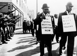 Black sanitation workers participate in a strike in Memphis in 1968 as National Guard members look on. One worker carries a sign that reads “End dismal working conditions now.” 