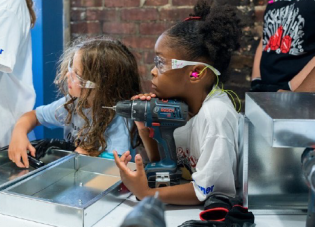 A young camper listens to an adult speak, her chin resting on a power drill, at Tools & Tiaras.