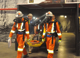 A mine rescue team wearing orange safety suits and respirators emerges from a tunnel carrying a stretcher after completing an exercise as part of the International Mine Rescue Competition.