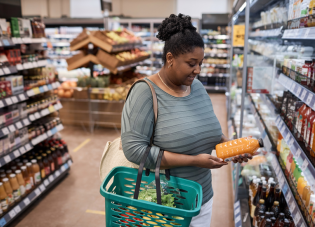 A woman holding a shopping basket looks at a bottle of juice in a grocery store refrigerator aisle. 