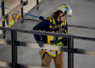 A woman construction worker wearing high visibility gear, a safety helmet with chinstrap and clear visor, a fall protection harness and gloves.