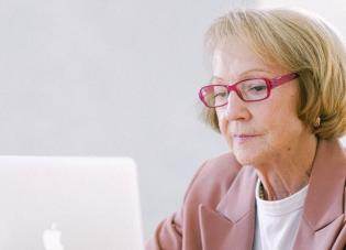 An older white woman with short blond hair is shown from the shoulders up. She wears red glasses and is looking at a laptop.