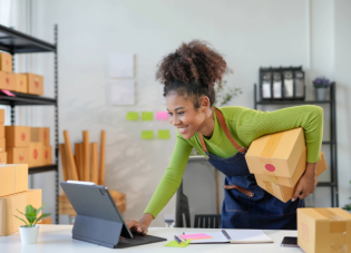 A young entrepreneur checks her laptop while holding shipping boxes.