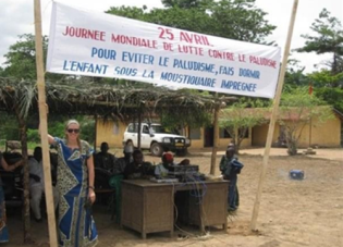 A young American woman in sunglasses stands by a group of people working behind desks in a dirt clearing. A sign in French advertises World Day Against Malaria. 