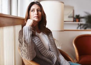 A woman sits in a living room with her head resting on her fist, looking pensively into the distance