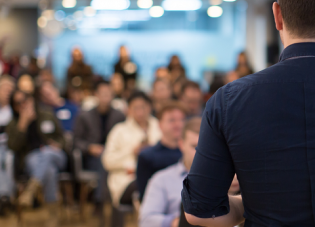 A stock photo showing the back of a male speaker as he addresses an audience 