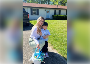 A woman crouches in a driveway to embrace a young boy. She holds an Easter basket full of colorful eggs.