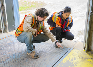 Two workers wearing high visibility jackets kneel down to examine a spill on the ground. 