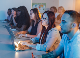 Students sit at a table in a classroom with computers and notepads in front of them to take notes.